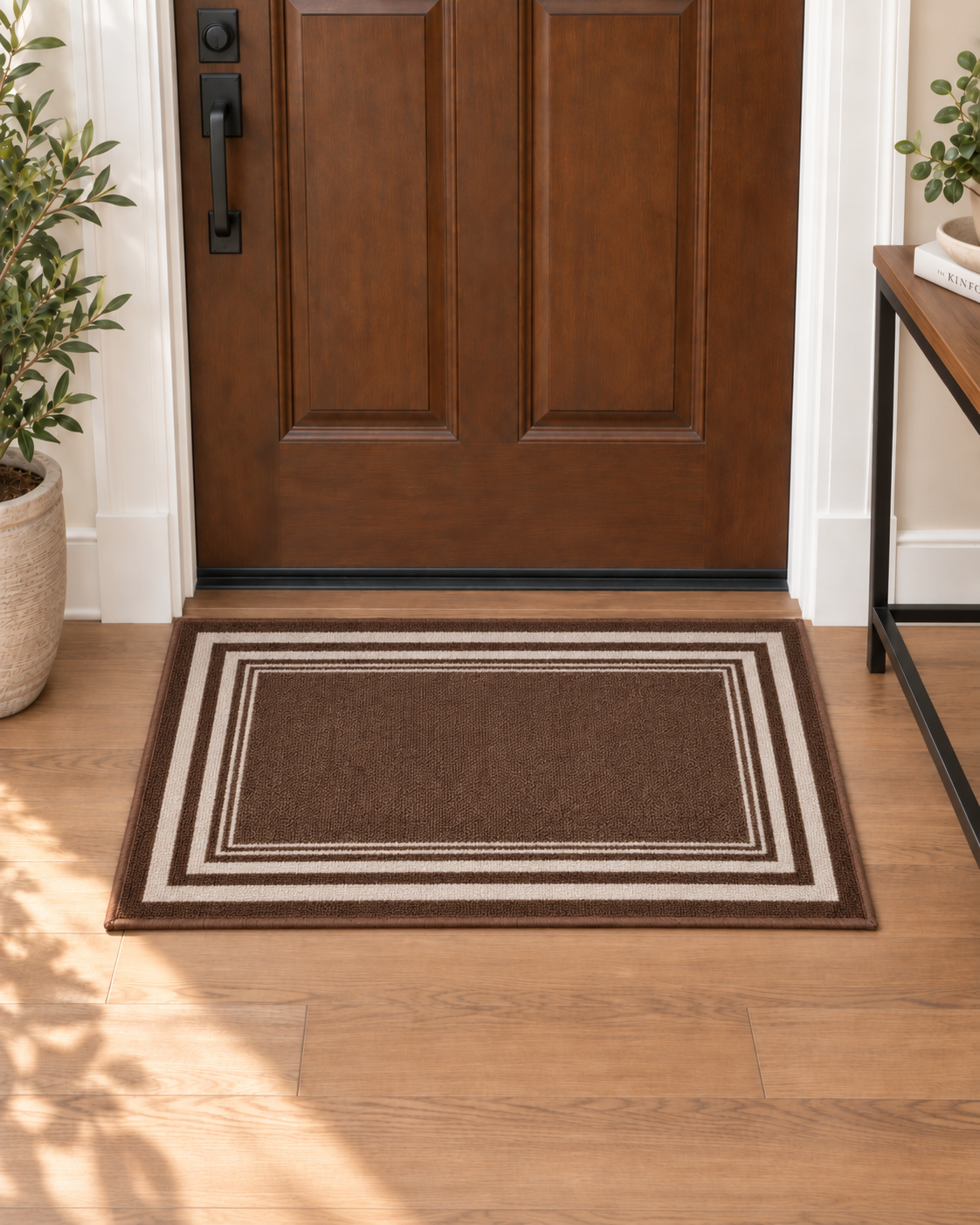 A rectangular brown doormat with concentric cream and brown borders, placed in front of a wooden front door on a light wood floor.